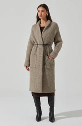 A woman is wearing a long, woolen, beige coat with a loose fit and large patch pockets on the front. The coat has a wide, draped collar and is cinched at the waist with a black drawstring. She is also wearing knee-high black leather boots.
